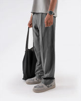 Person wearing grey straight pleated trouser and shoes holding a black bag on a white background
