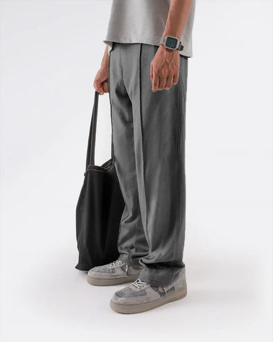 Person wearing grey straight pleated trouser and shoes holding a black bag on a white background