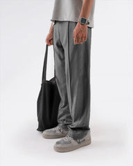 Person wearing grey straight pleated trouser and shoes holding a black bag on a white background